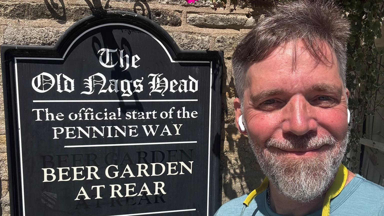 Mr Neal takes a selfie in front of a sign for a pub which also signals the start of the Pennine Way. He has grey hair and a grey beard. He wears a blue T-shirt with earpods in.