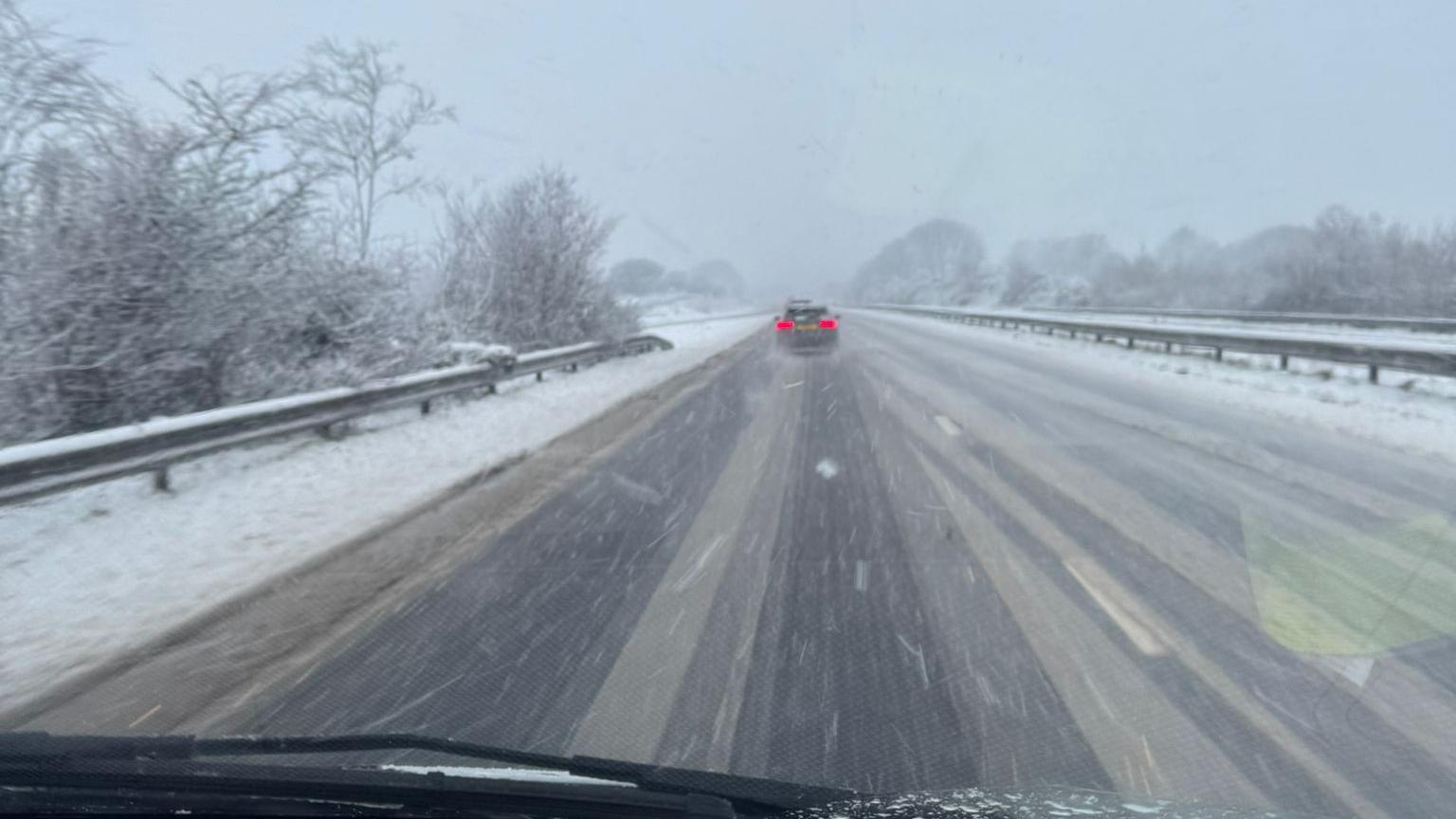 Snow falls on the A30 in Cornwall near the Jamaica Inn pub. The road has a light scattering of snow on it with tracks from where vehicles have driven clear to see. A car is in the distance with its brake lights on.