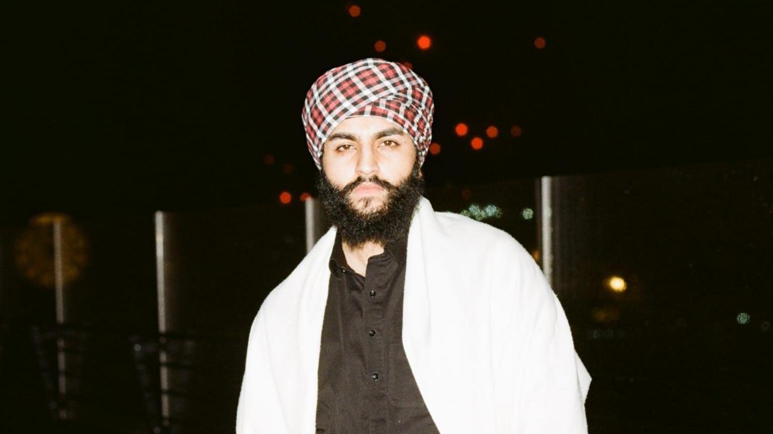 Beant wears a red and white checkered turban with a black top and white shawl.