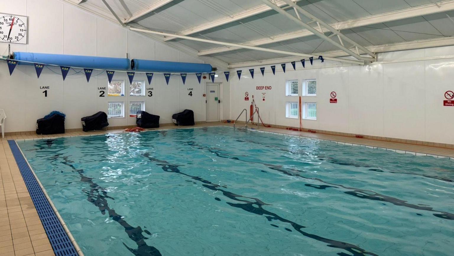 The photo shows a swimming pool. Above it, spanning the length of it is some blue bunting. There is a clock displaying the temperature to the left hand side of the photo. There are also stickers marking each lane on the wall. 