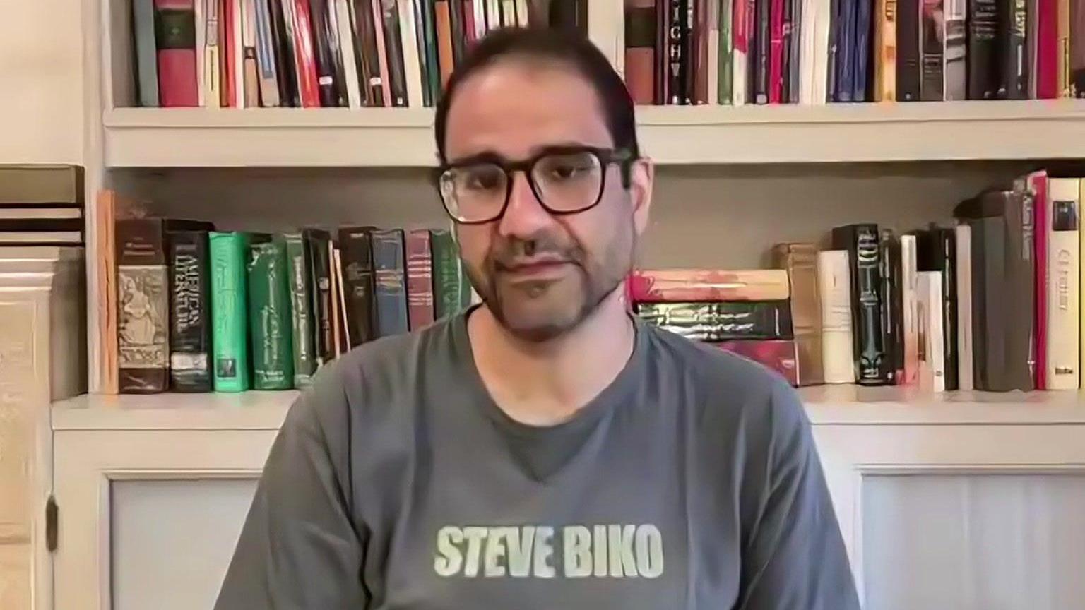 Alaa Abdel Fattah speaks via video link to the BBC, sitting in front of a bookshelf and wearing a grey t-shirt with the words Steve Biko on it