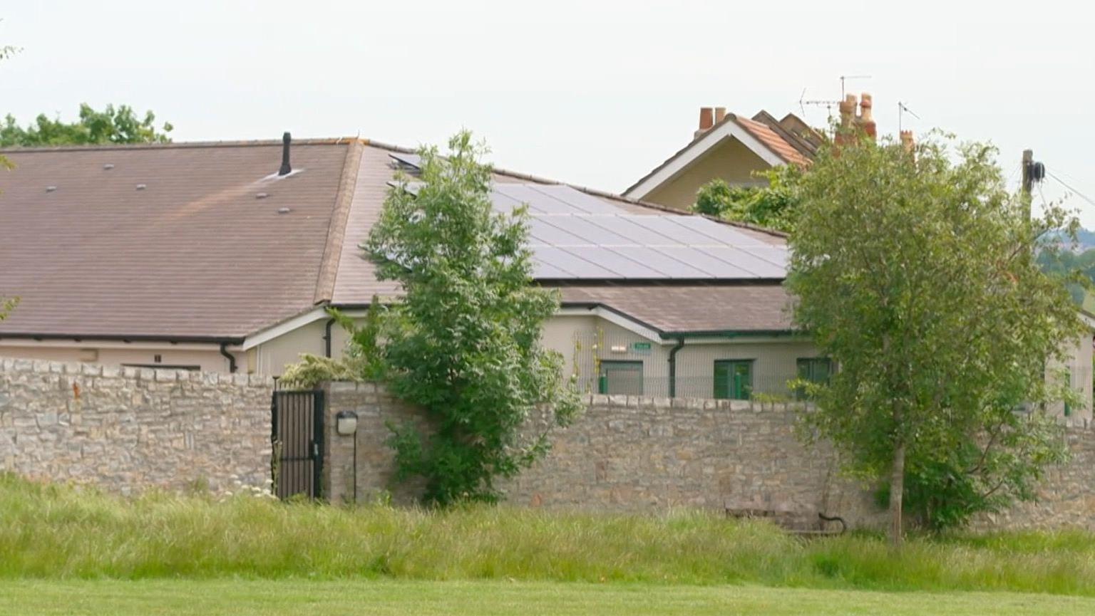 The exterior of a large building inside a tall stone wall. The building has a sloping roof of light red tiles and black guttering. There are some trees visible.