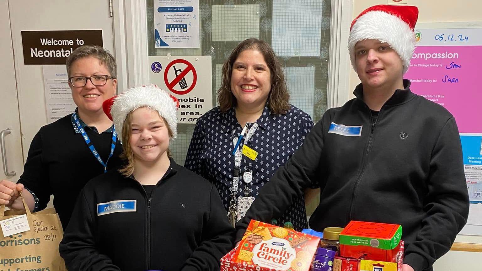 A woman in a black jacket holds a paper bag in a hospital ward. She stands next to a woman and a man wearing Santa hats. The man is holding a open hamper of biscuits, tea and snacks. A woman in a navy dress stands between them smiling at the camera.