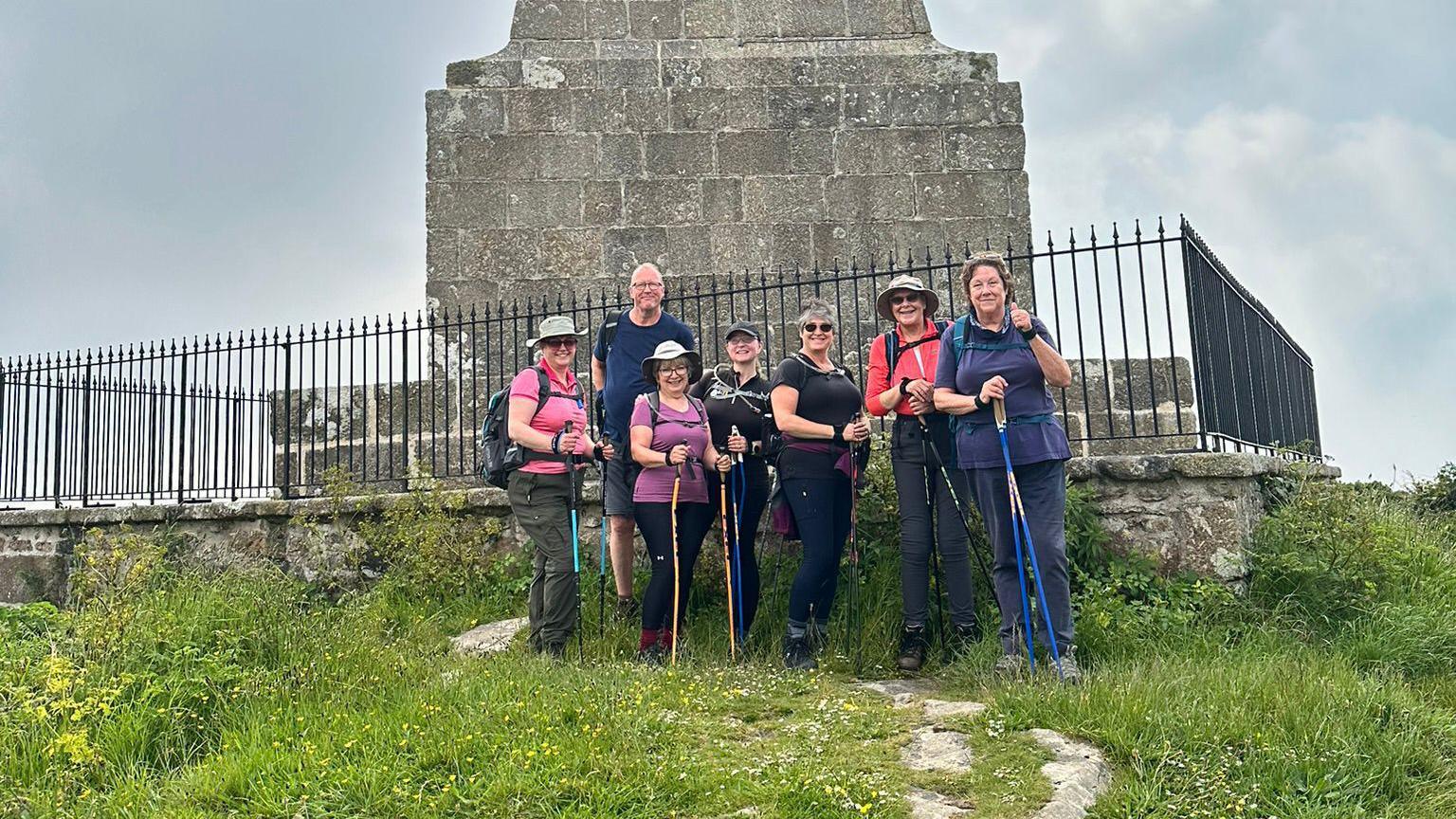 Seven members of Walk Kernow stand in front of Knill's Monument on on St Michael’s Way in Cornwall. All the members have walking poles. It is a cloudy day. The granite monument has a black metal fence around it.