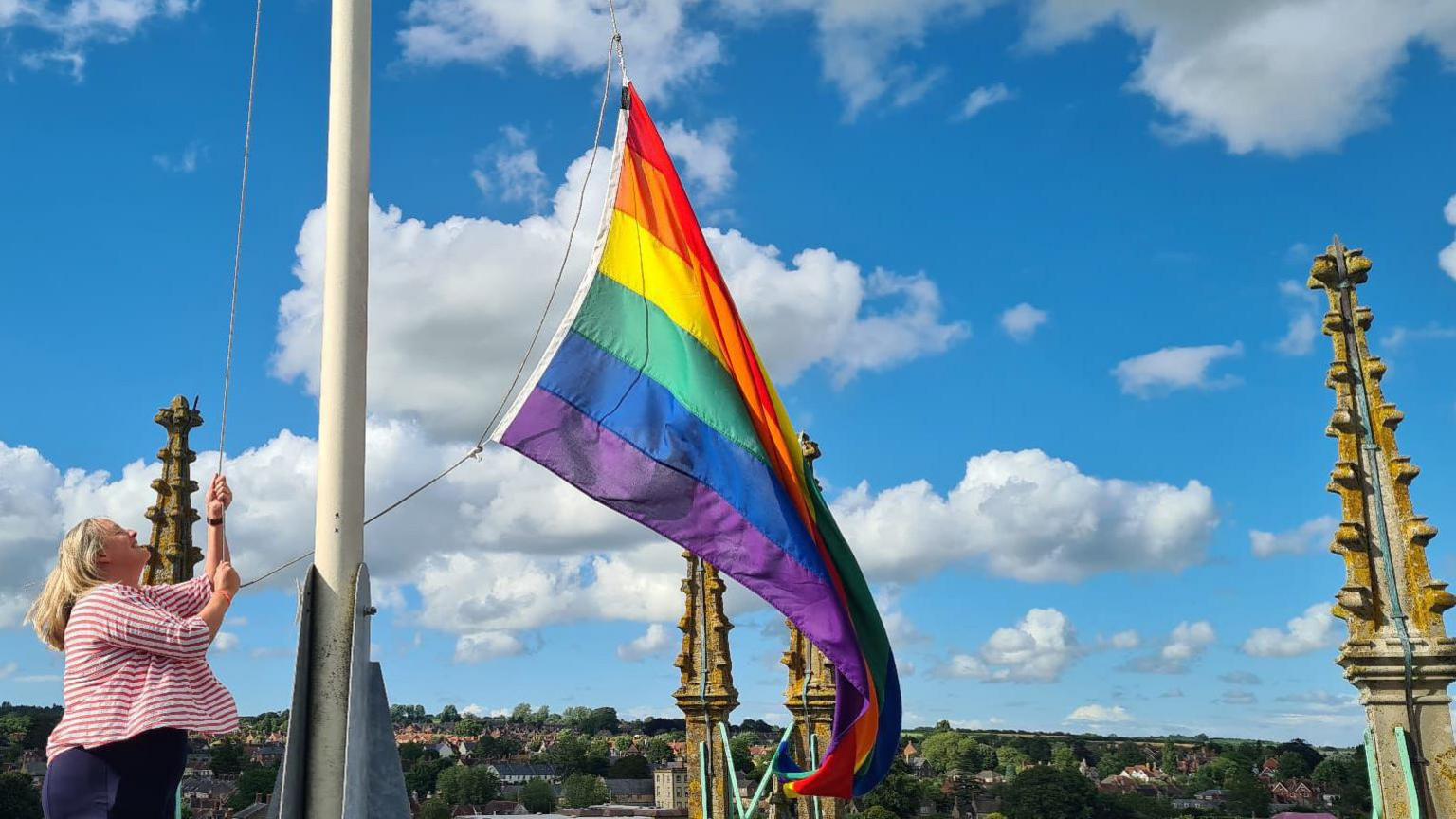 A woman raises a Pride flag on the mast above Sherborne Abbey, the town skyline behind her, a blue sky with clouds above