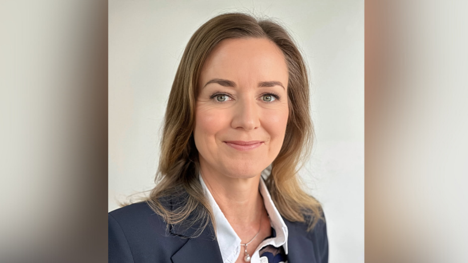 Elizabeth Stephens smiling with shoulder length brown hair, a blue suit jacket and a white blouse. She has a necklace with a pearl pendant. There is a plain beige background.