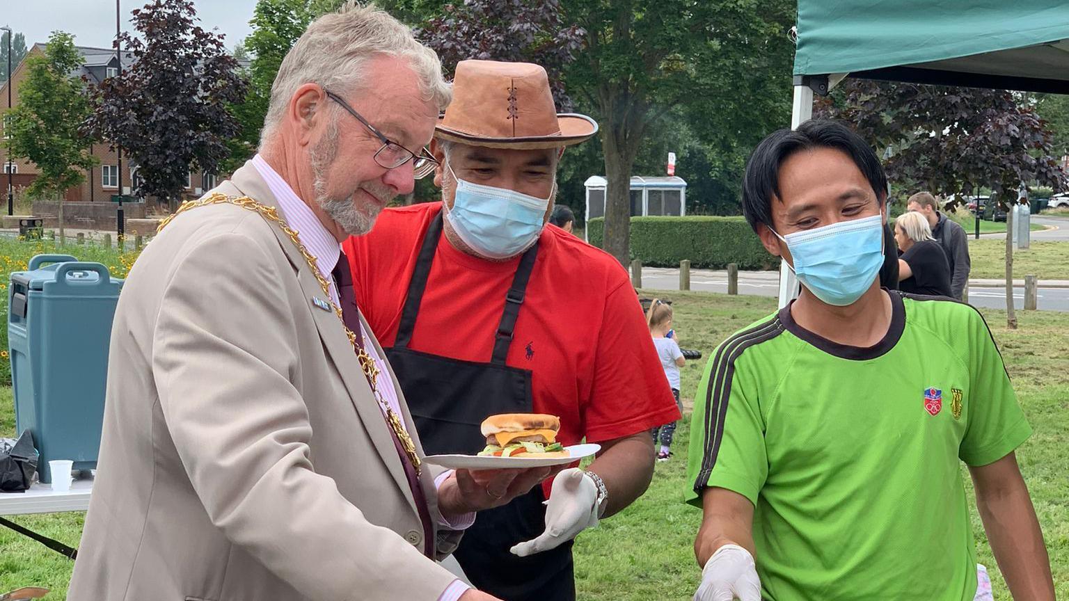 A man with short grey hair and a grey beard and glasses stands holding a plate of food. He is wearing a light brown or grey suit over a pink pinstripe shirt and a burgundy tie. Two other men are nearby wearing facemasks. One has black hair in a centre parting and is wearing a green t-shirt. The other is wearing a black apron over a red t-shirt, and a hat.