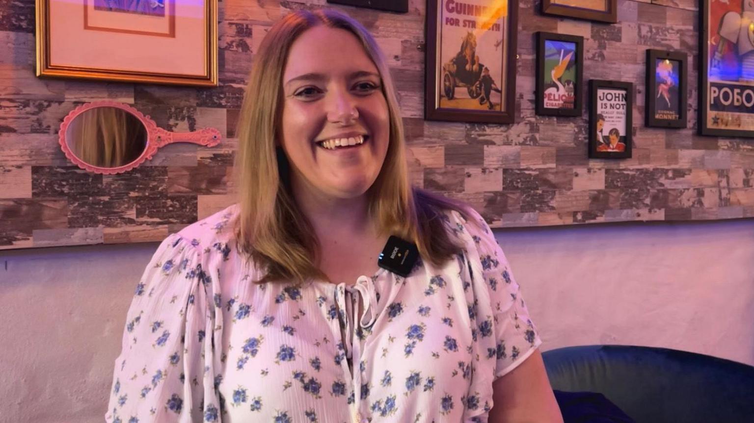 Poppy Murray is pictured. She has shoulder-length light brown hair and is wearing a short-sleeved white top with a floral pattern. Behind her is a wall which is covered with framed posters.