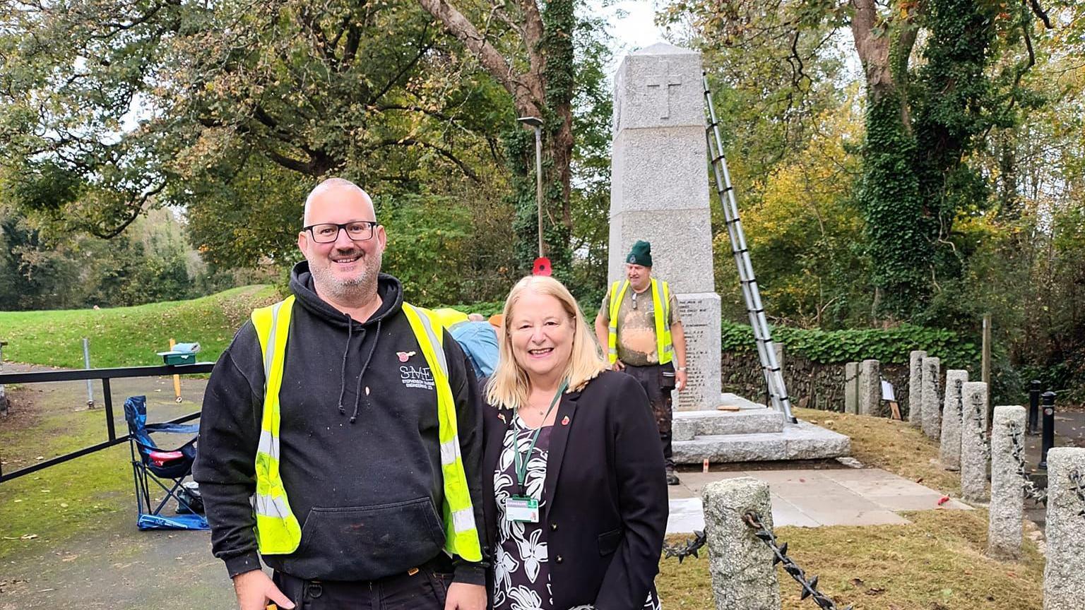 Andrew Stephenson on the left wearing a black hooded jumper with a hi-vis over the top and Terri Beer stood next to him wearing a black blazer and a poppy badge on the right. They are both smiling at the camera. In the background is Steve Davies who is working on the war memorial which has a ladder leaning against it.