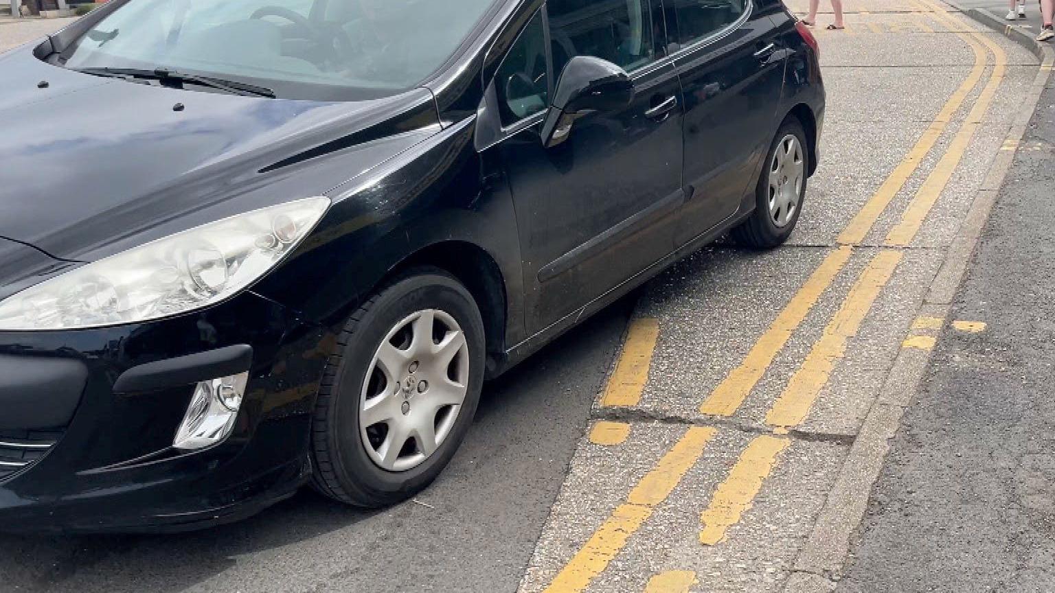 A black car pulls away after stopping on double yellow lines at the Sea Lane bus stop