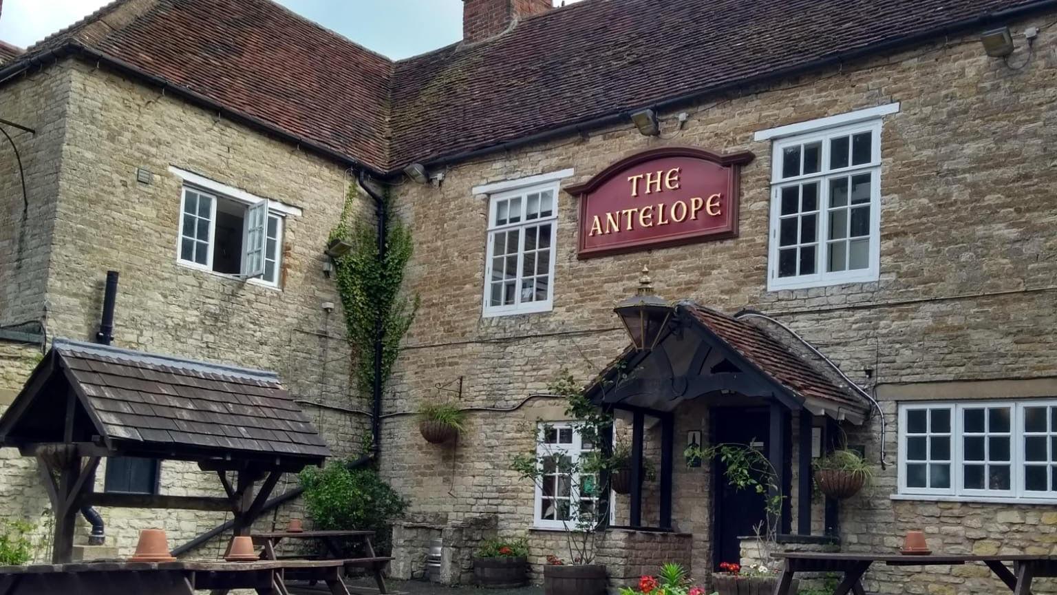 A traditional country pub built in honey-coloured stone with a red name sign 