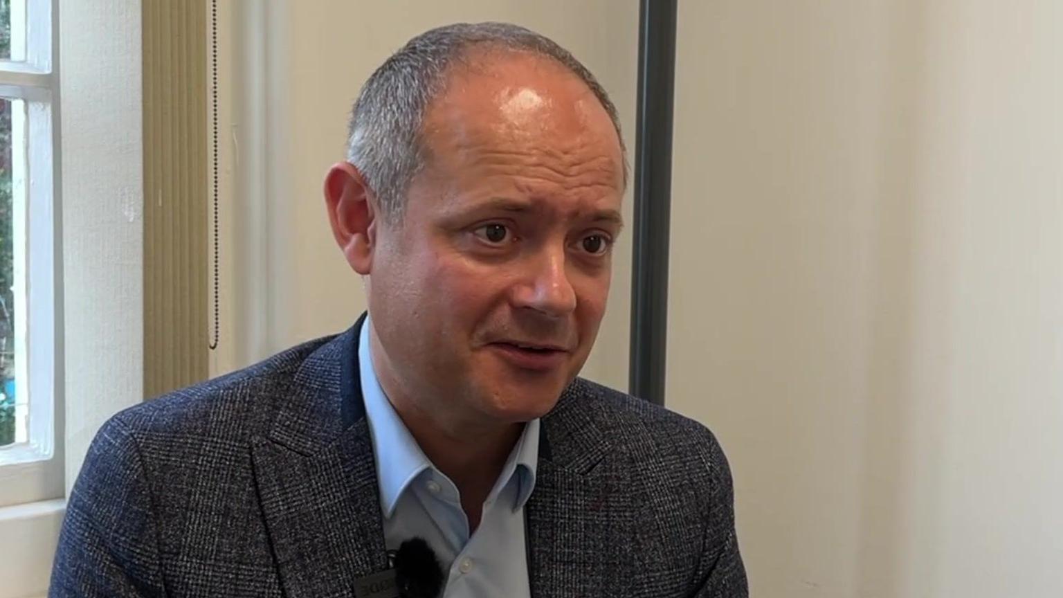 Steve Harris wears a grey jacket and light blue shirt in a still from a TV interview. He has short, grey hair, balding at the front.