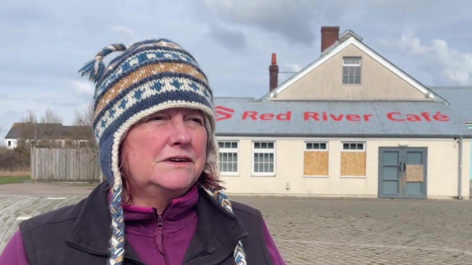 Dawn Harries is an older woman wearing a multi-coloured woolly hat, a purple fleece and a brown gilet. She is standing in front of a boarded-up cafe in Heartlands.