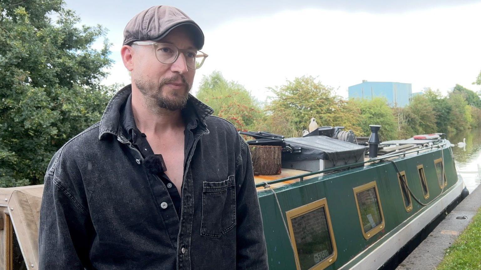Robbie Cumming standing in front of his canal boat. He is wearing a cap, glasses and has a trimmed beard.