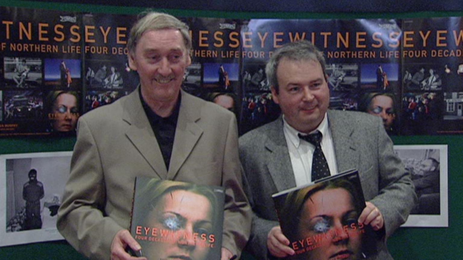 Brendan Murphy wearing a tan blazer and black shirt holding a book with a woman with shattered glass over one eye which is titled Eyewitness. Beside him in Seamus Kelters wearing a grey blazer, white shirt and black tie holding the same book.