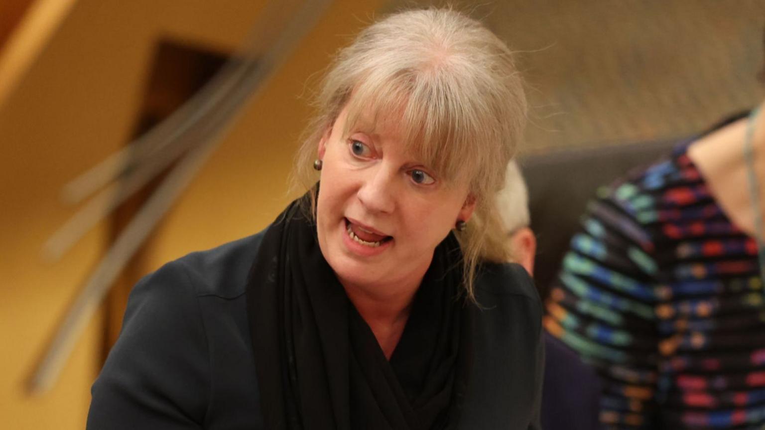 Shona Robison looks intently at a colleague in the Holyrood chamber while delivering her budget speech. She is wearing a black blazer and has blonde hair tied back with a fringe