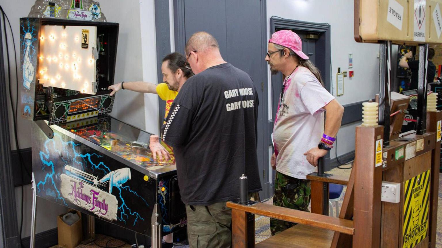 Three men moving an Addams Family pinball machine into position. There is a wooden chair which looks like an electric chair in front of it