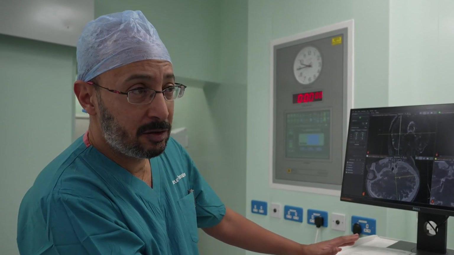 Jibril Farah: a bearded man, wearing a blue surgical outfit and head wrap, pointing to a brain scan on a PC monitor.