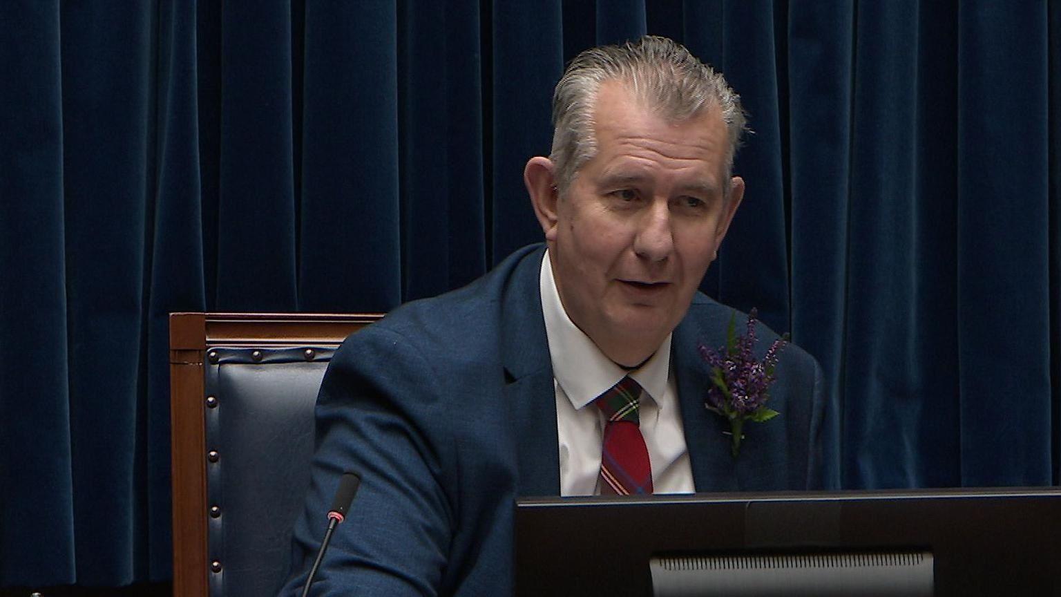 Edwin Poots in the Speaker's seat in Stormont. He has grey hair and wearing a dark blue blazer, white shirt, red tartan tie and buttonhole floral decoration of purple heather.