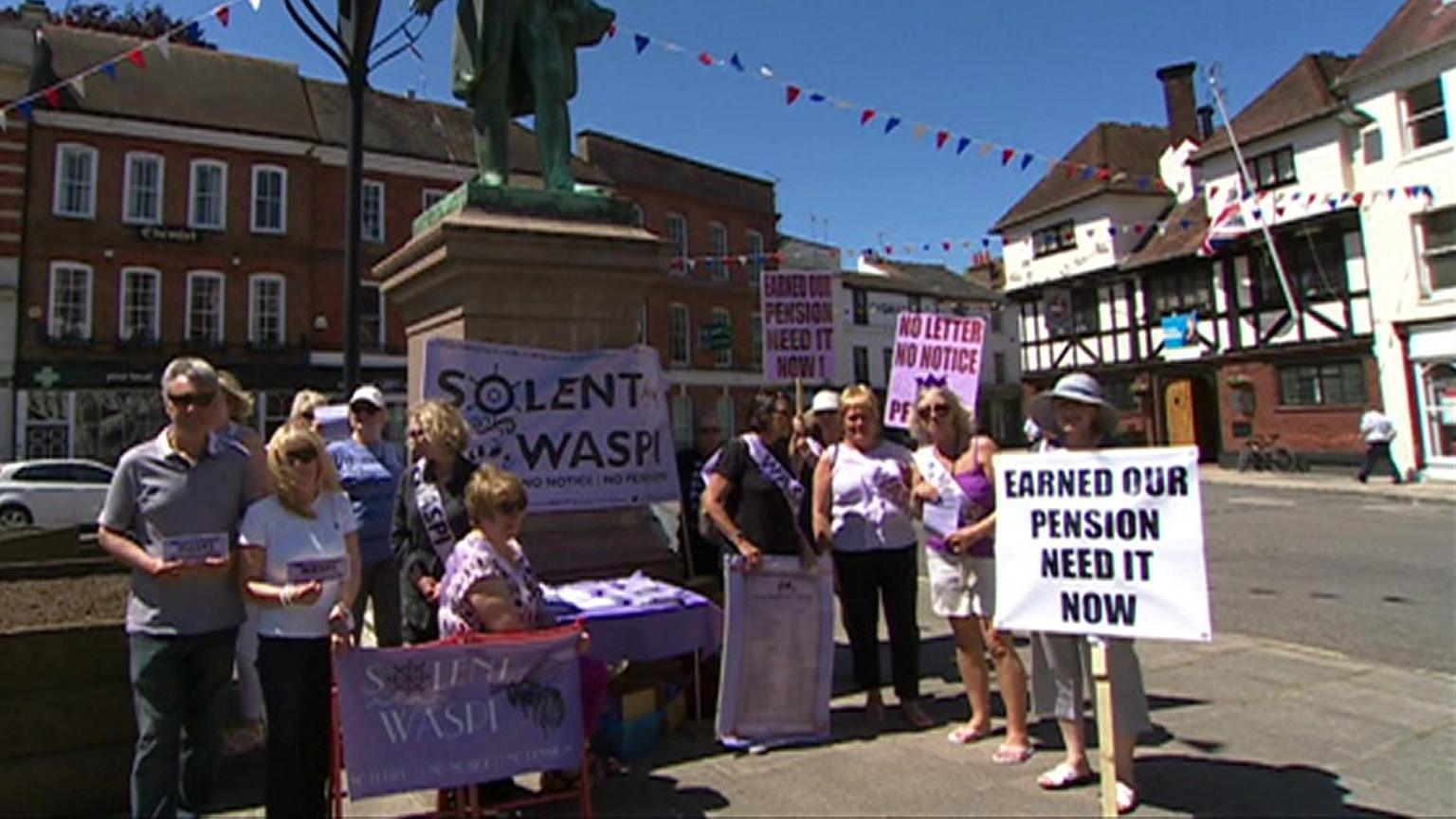 A group of women and one man holding banners saying "earned our pension need it now" and "Solent WASPI" and "no letter, no notice". They are in Romsey market square in front of the statue of Lord Palmerston. It's a sunny day in 2017. 