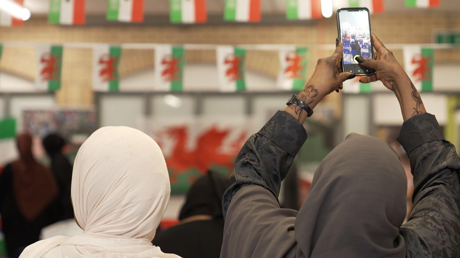 Two women in head scarves are seated facing away from the camera, the one on the right is holding up a camera to take a picture of Welsh and Somaliland flags hanging in a conference room.
