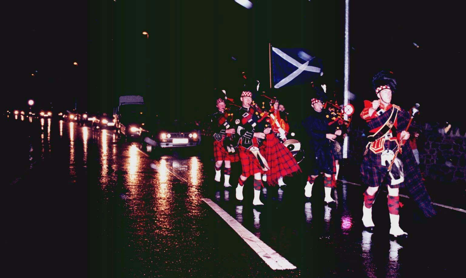 A group of bagpiers in Scottish dress and carrying a Saltire walking along a road after dark. A line of car headlights can be seen behind them.