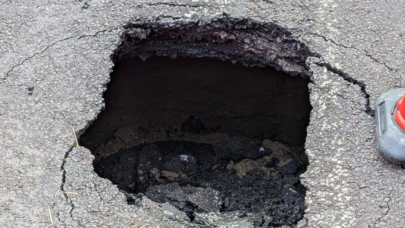 A close up of the sinkhole shows the collapsed road surface inside the hole surrounded by cracked tarmac and a traffic cone.