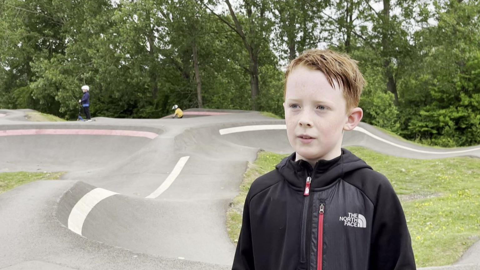 Ashton is a young boy with red hair. He is standing in front of Chopwell pump track. A boy in a scooter is standing on the track behind him, waiting his turn while another boy is riding a bike on a corner.