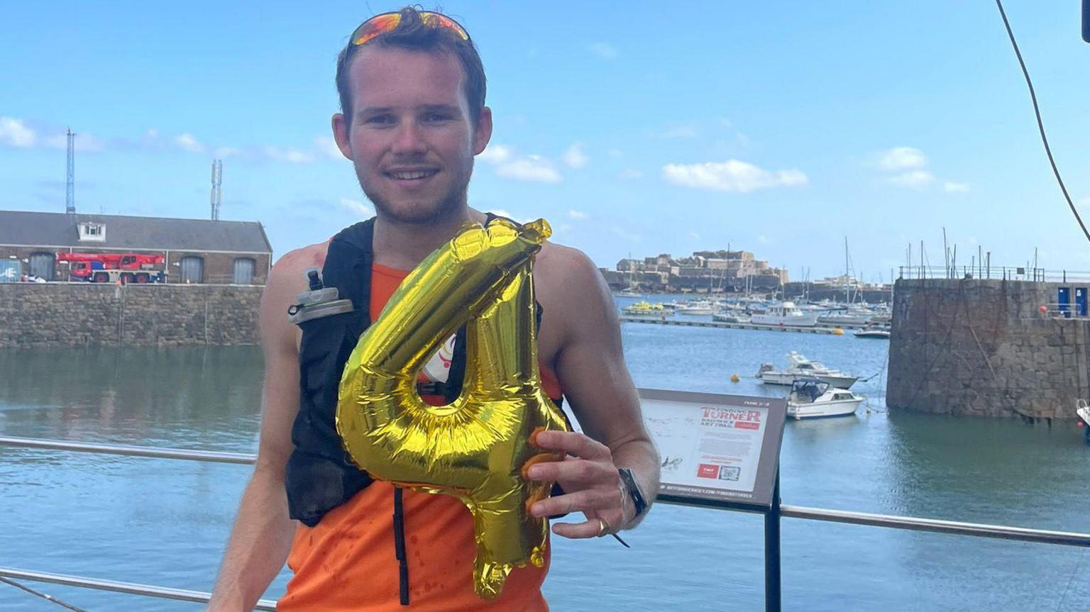 James stands near a waterfront holding a large gold balloon shaped like the number '4'. They wear an orange sleeveless top, black shorts, and a hydration pack. Behind them, boats are docked and buildings line the shore under a clear blue sky