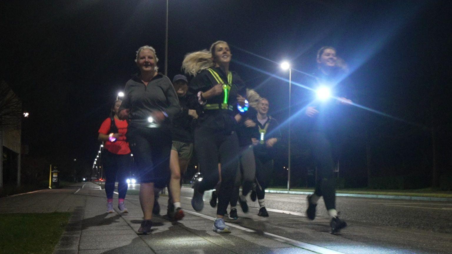 A group of women run on a street at night. There are seven women on a pavement. They are wearing lights and reflective tops. Behind there is a street light.