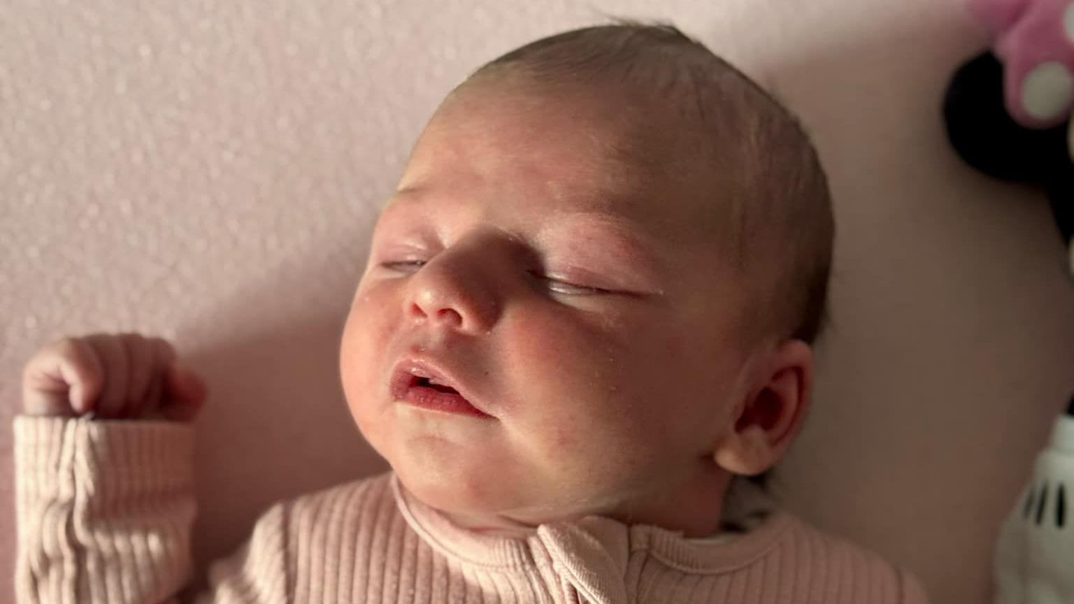 A new born baby lying in a cot next to her teddy bear. It is a close up shop and the baby is wearing a baby pink outfit.