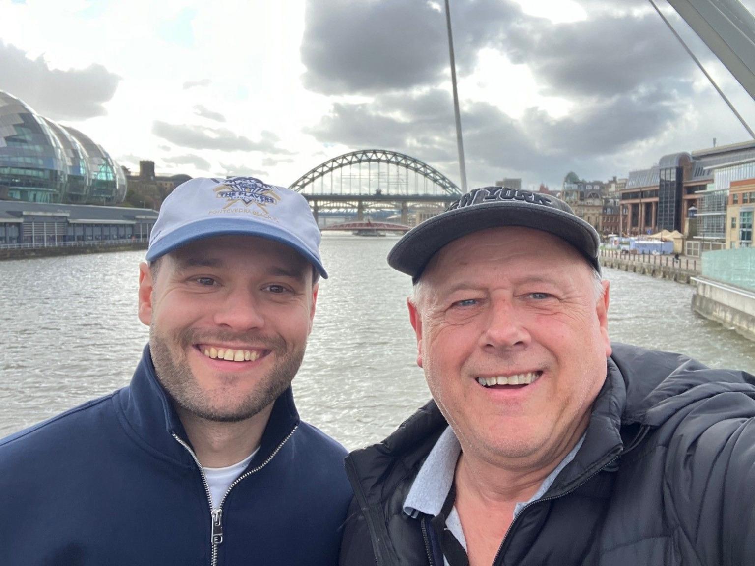 Kieran is smiling at the camera. He is wearing a dark blue fleece jacket and cap. His dad, Brian, is also wearing a cap and puffa jacket. The Newcastle bridge and Tyne River are in the background.