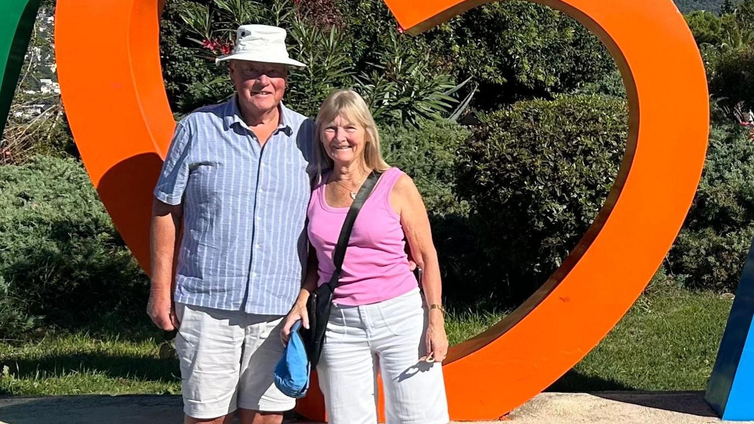 A man in a white hat and striped shirt next to a woman in a pink top with an orange heart shape behind them.