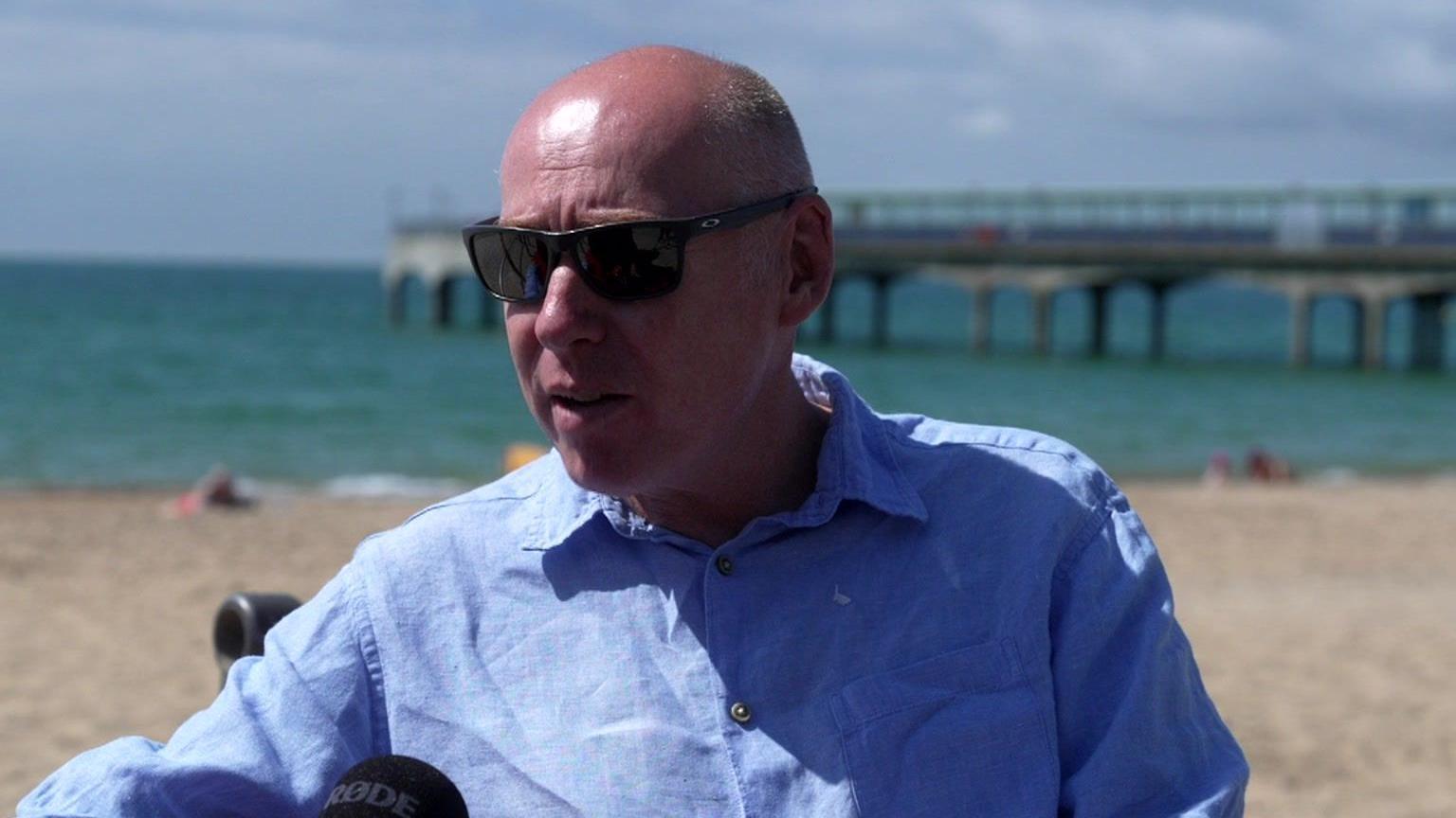 Tony is wearing a light blue shirt and sunglasses while sitting in front of Bournemouth beach