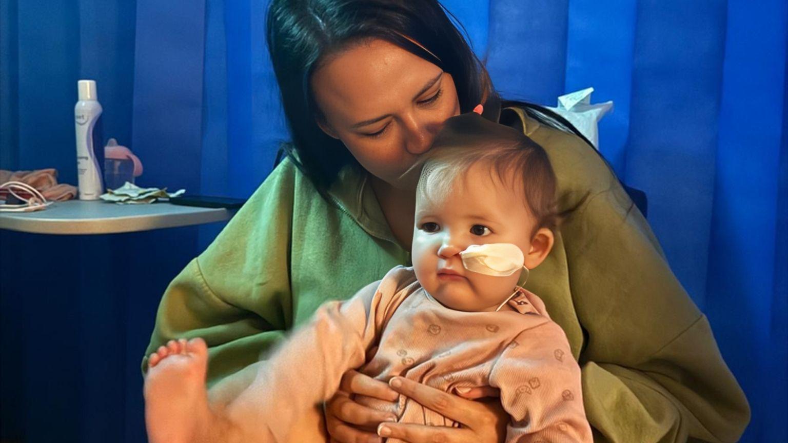 Olivia Brummitt and her baby daughter Winnie sit cuddling in a hospital room. There is a blue curtain in the background. Olivia is wearing a green top and Winnie is wearing a pink baby grow.