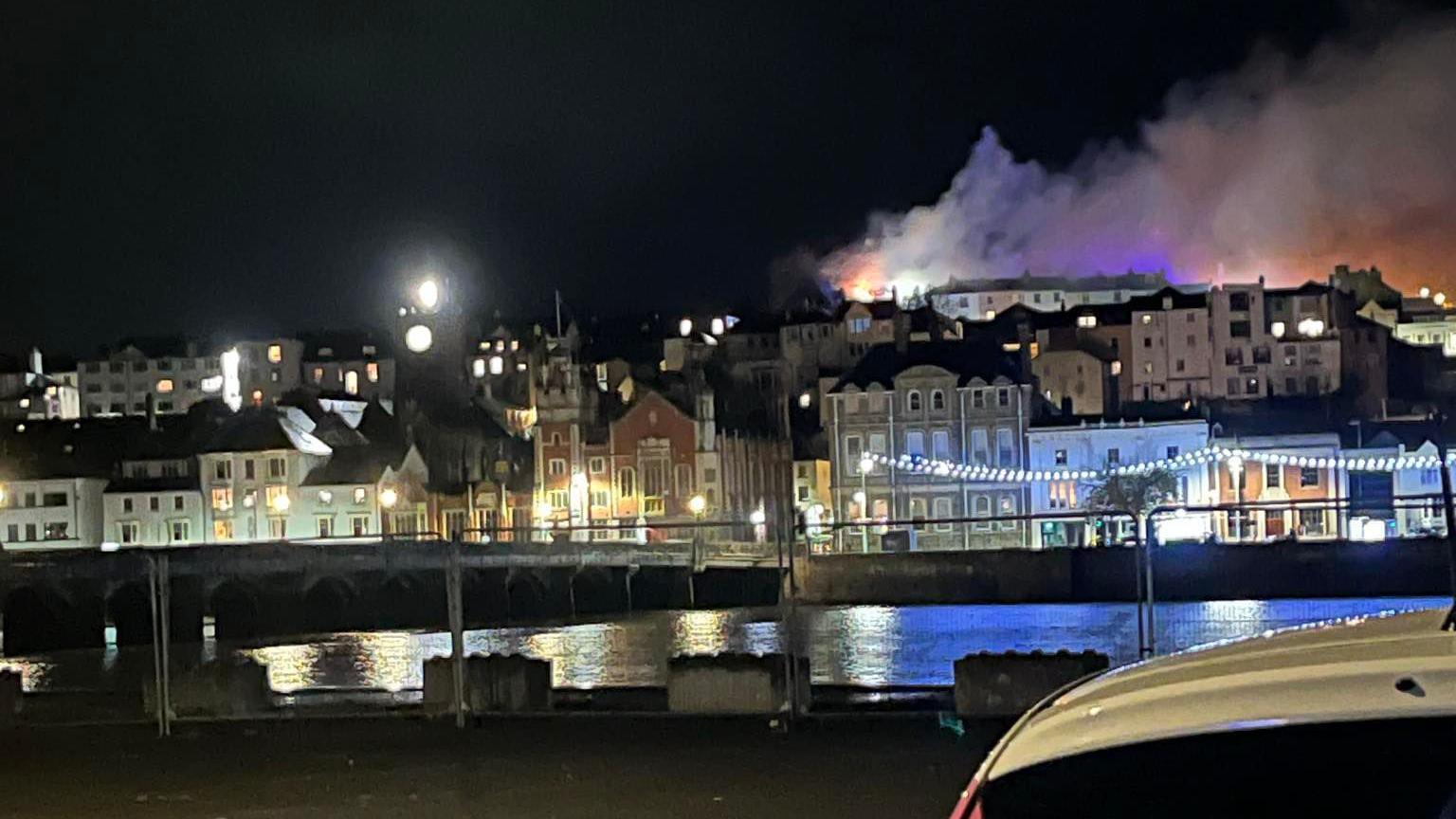 Long range shot of a fire sending smoke and flames into the air into the night sky over Bideford. Dozens of buildings with their lights on are on the opposite side of the river from where the photographer is stood. A bridge connecting the two banks is on the left-hand side.