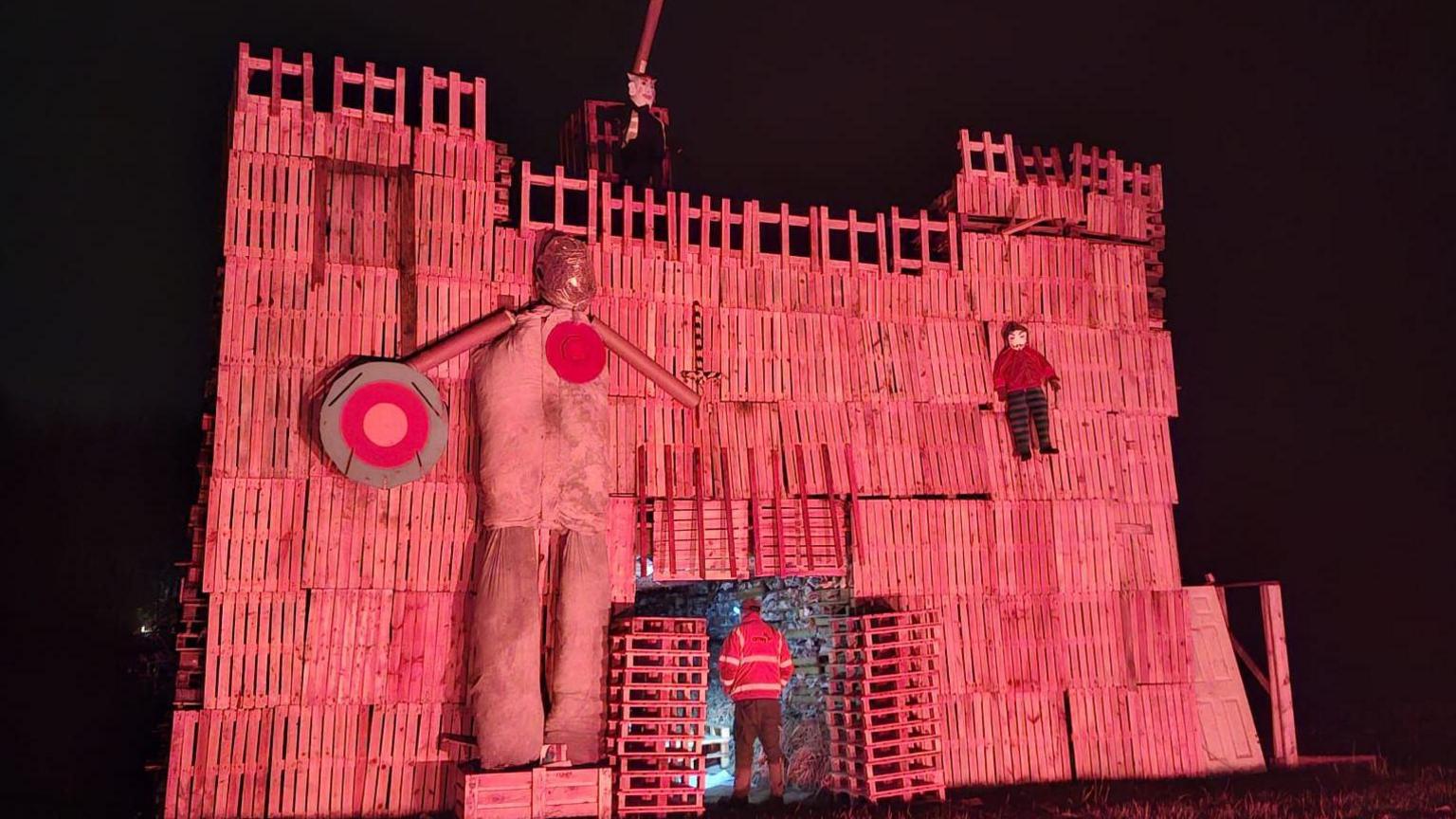 A fort shaped structure at least two storeys high made out of old pallets, with turrets and scarecrows. It is night time and the structure is light with a pinkish-red light.