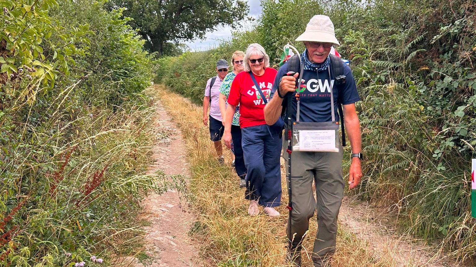 John leads the way down a narrow country lane, with tall hedges either side. He holds a walking stick, with his family members walking in a line behind him.