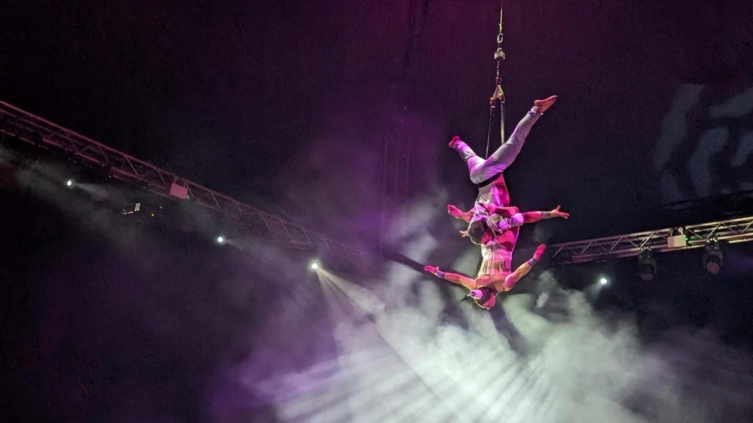 Two circus artists are hanging from the ceiling on a rope. Their arms are stretching out. Lights are pointing at them.