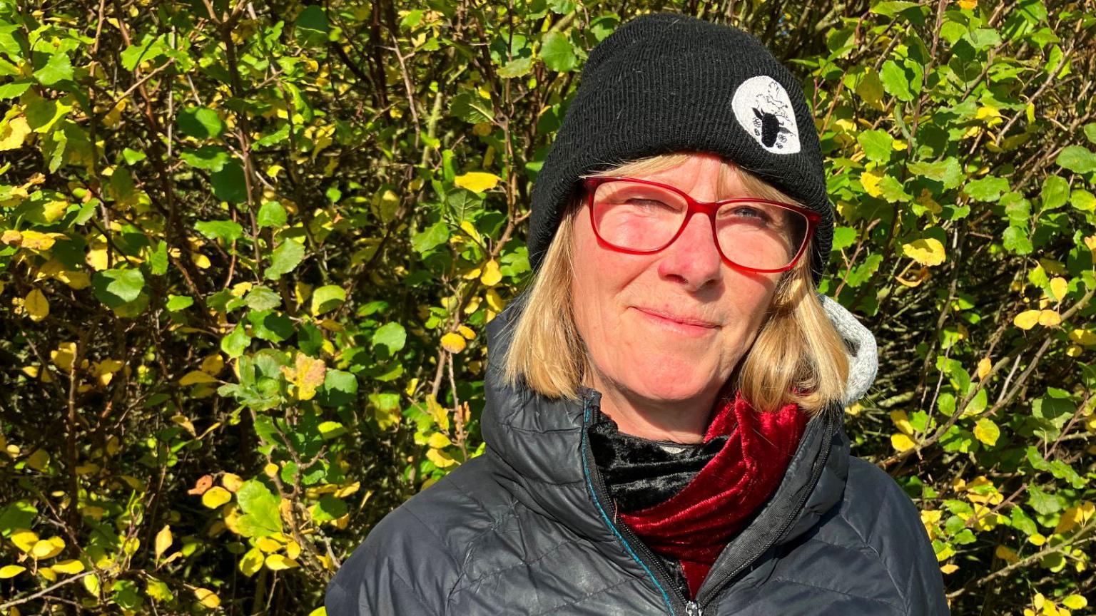 Kathy Barley, who has a blonde bob and wears a black hat, stands in the foreground, with a hedgerow in the background. 