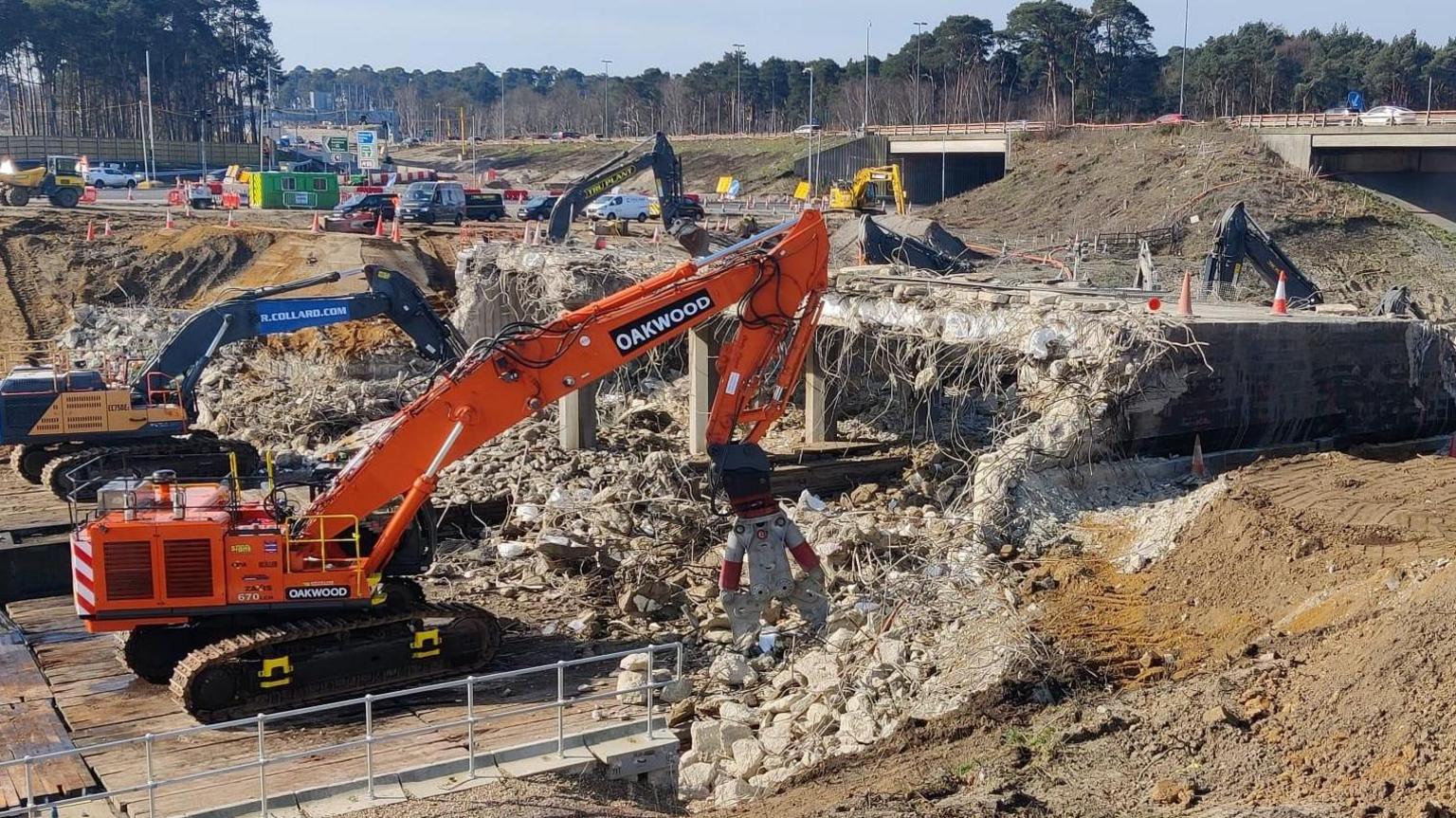 A yellow digger works to clear rubble from beneath a motorway bridge which has been partially demolished.