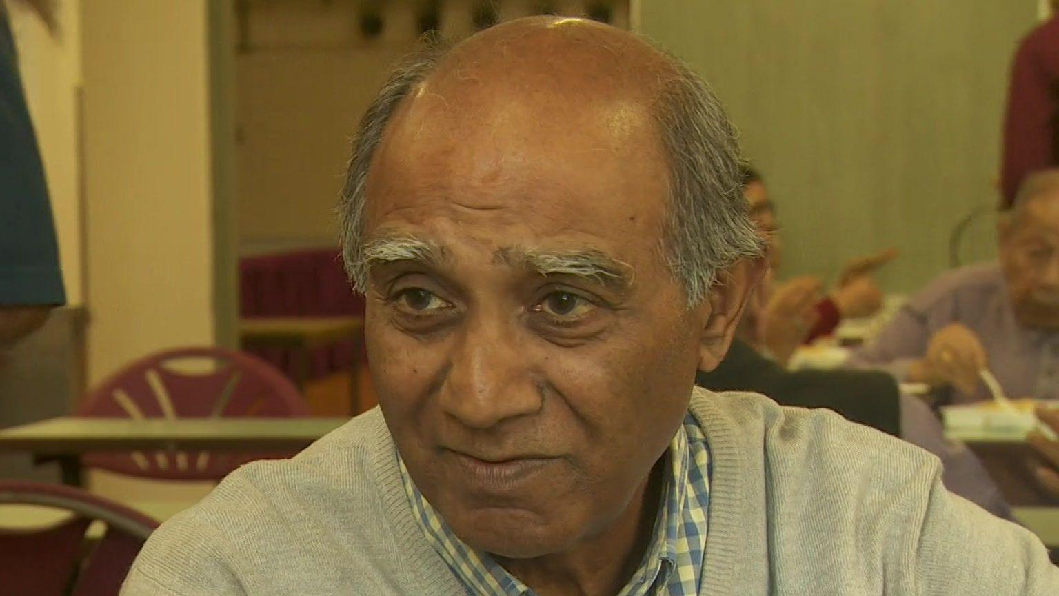 Ishwer Tailor with balding, grey and dark hair, wears a blue and white checked shirt with a grey sweatshirt, and is looking to the left of the camera as he sits in a chair with a room full of people in a restaurant setting.