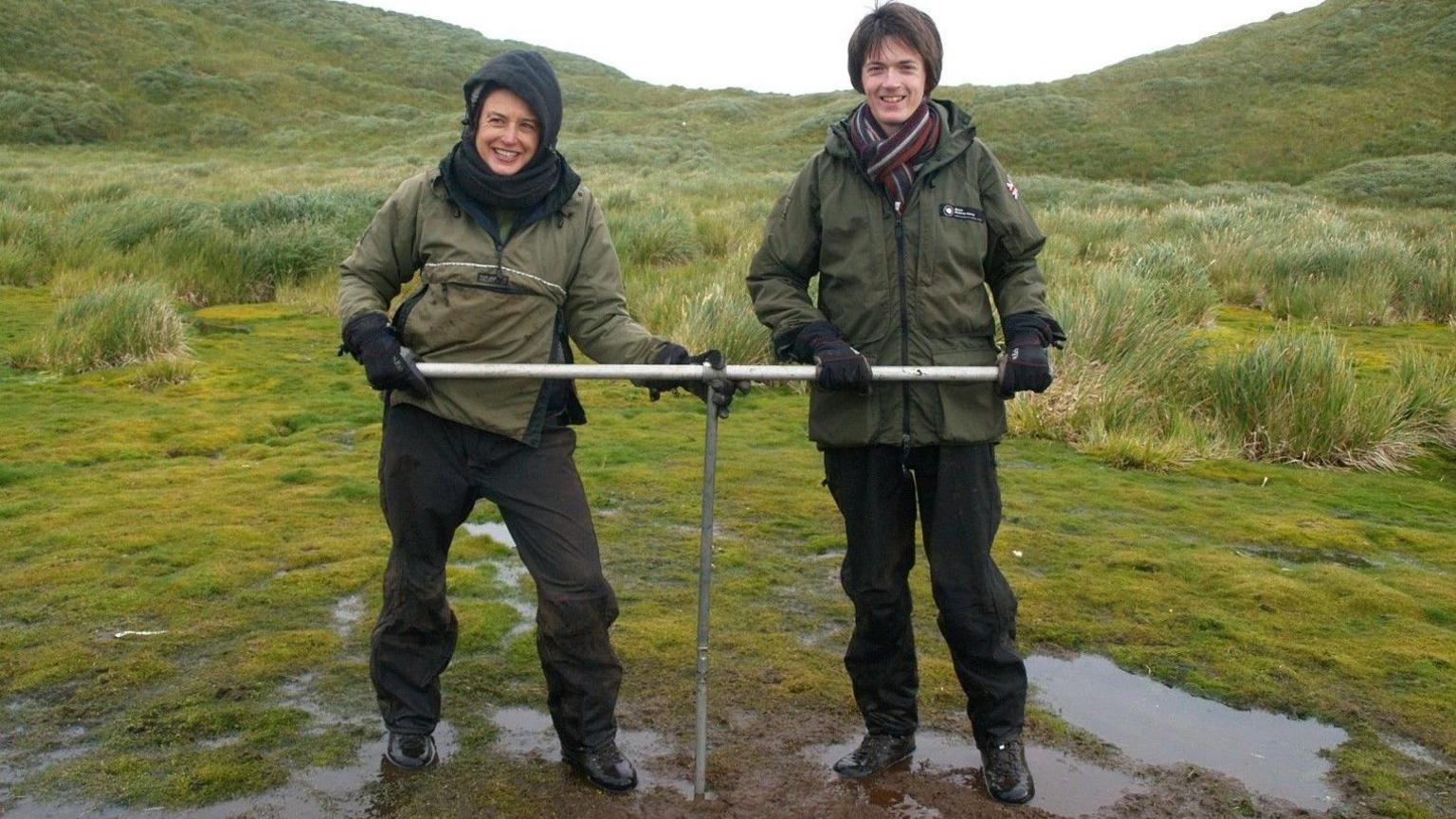 Two scientists are shown in wet weahter gear holding a piece of equipment used for collecting peat cores on Bird Island.