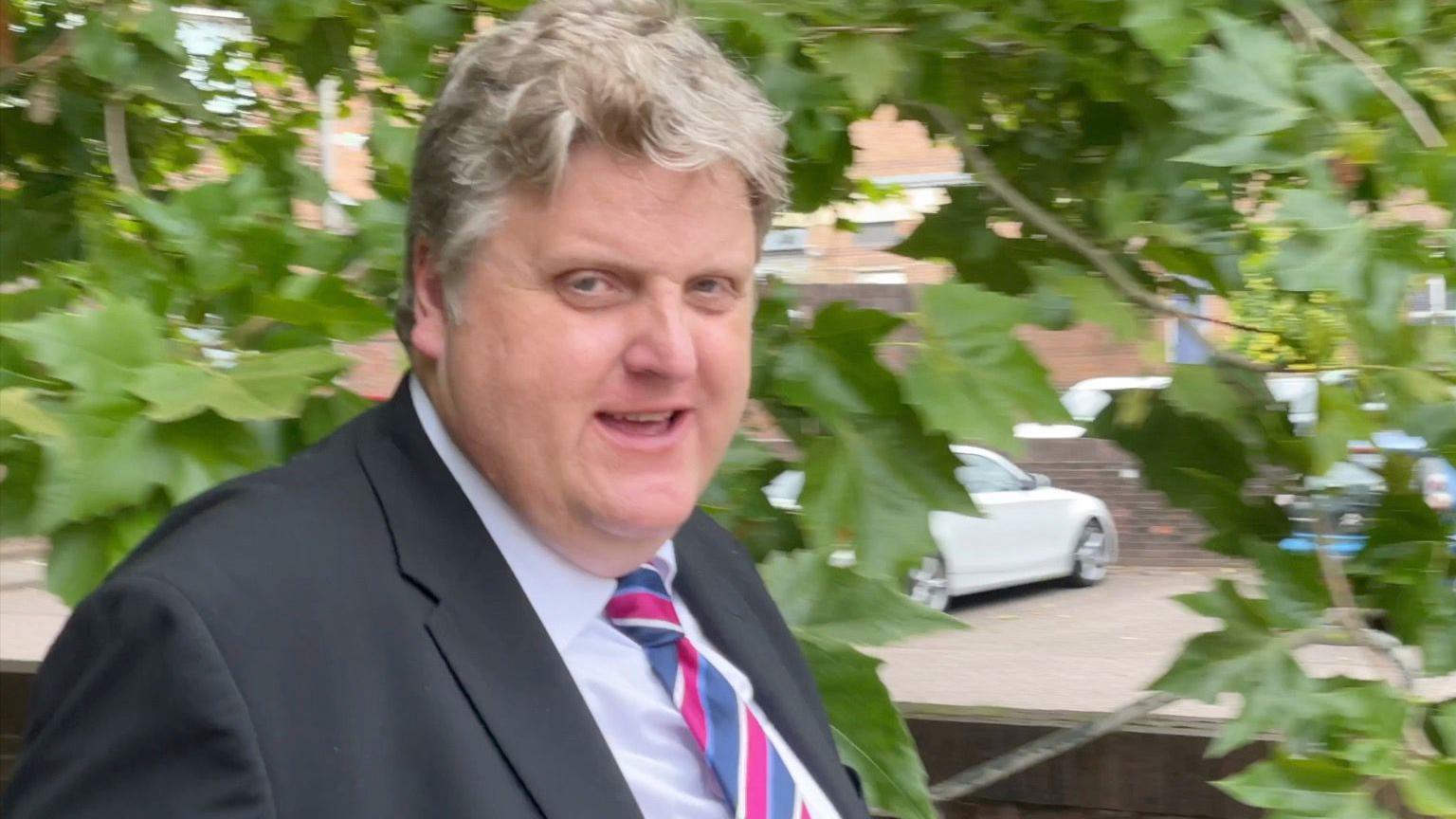 Gregory Hill photographed as he walks quickly away from the camera, passing a tree. He is wearing a suit and multi-coloured tie. 