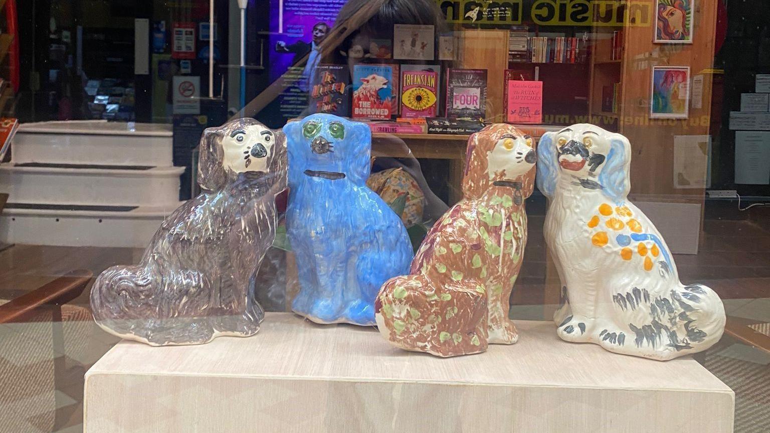 Four pot dogs in a bookshop window. One is painted black, one blue, one brown with green spots and one is white with yellow spots and grey and blue markings 