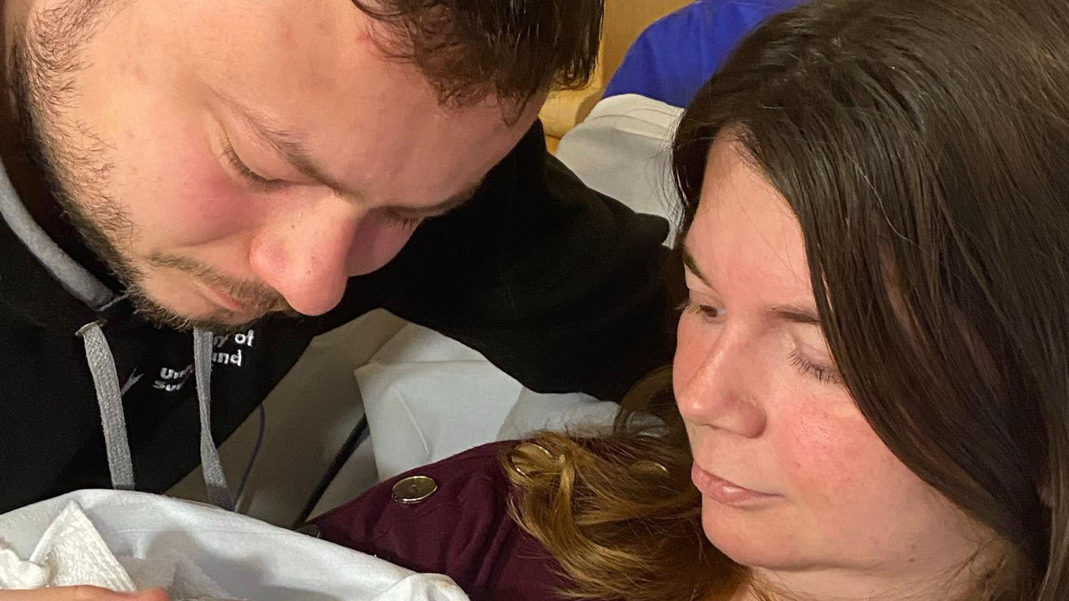 Alistair and Amy Peel are holding their heads close together and looking off camera to baby Lucie-Mae. Mr Peel has brown hair and a beard. Mrs Peel has long brown hair and is wearing a maroon top.