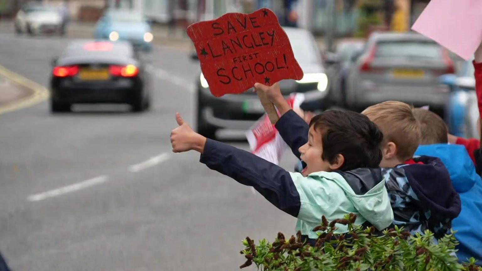 A group of young boys facing cars on the road with placards. One boy is holding up a handmade sign which reads 'Save Langley First School' in one hand and holding his thumb up with his other.