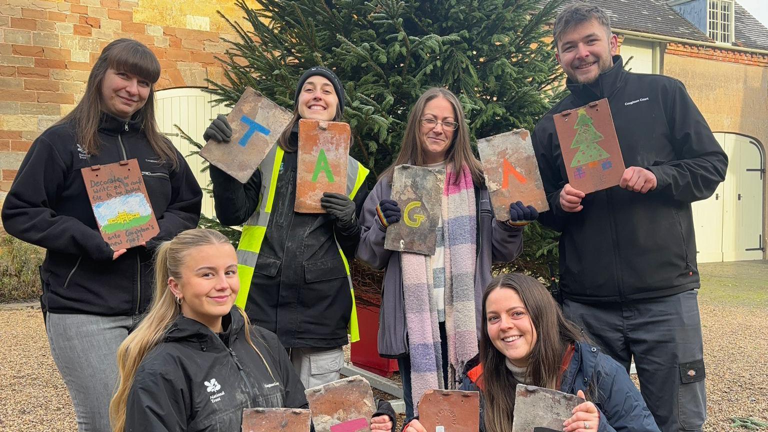 Six people gather in front of a Christmas tree on a gravelled courtyard and hold up tiles with images and words written on them.