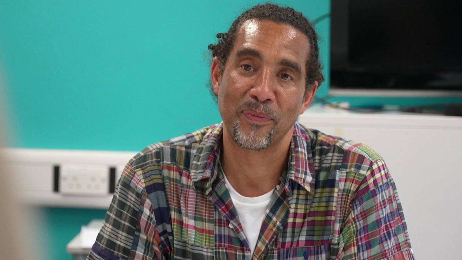 Paul Anderson sits at the table taking part in the discussion, a bright blue wall behind him. He has his hair tied back and wears a colourful plaid shirt