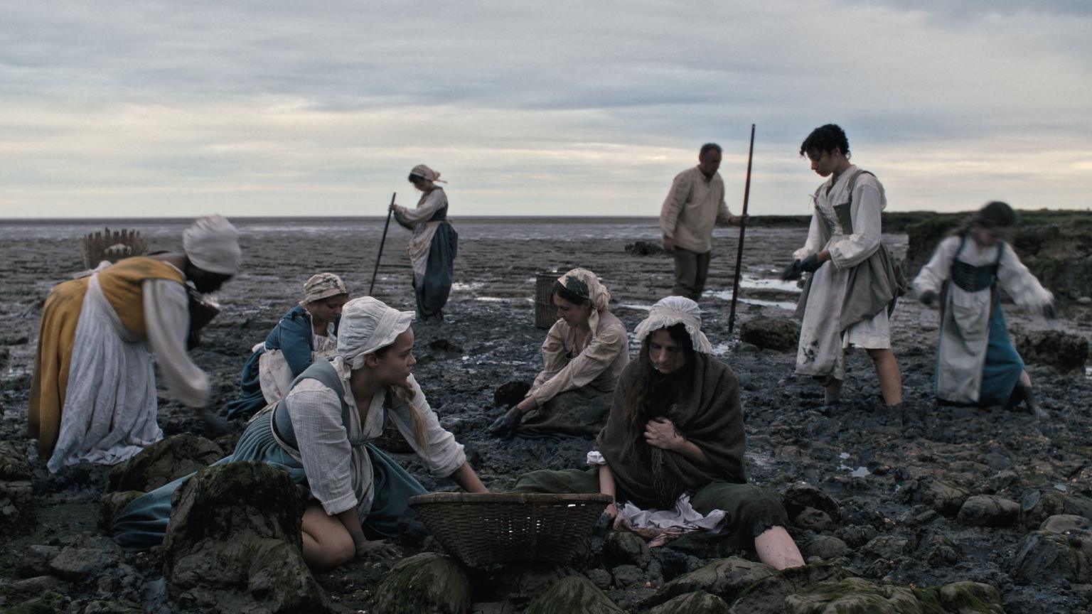 A still image from the Gossip film. A group of women sit and stand on a beach. They wear dresses, aprons and hats from the 16th Century.